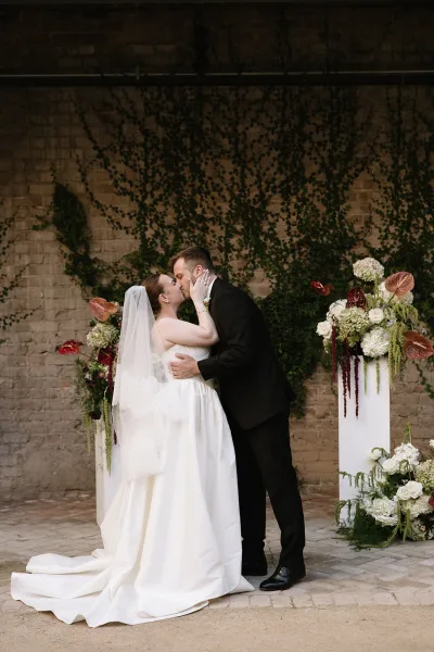 Ceremony kiss moment as the wedding first kiss is shared before an ivy-clad brick wall, the bride's veil floating while they embrace.