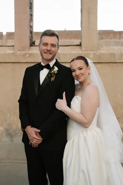 Couple portrait with bride holding the groom's arm, her lace-trimmed veil catching light, set against a stone wall with window openings and sky.