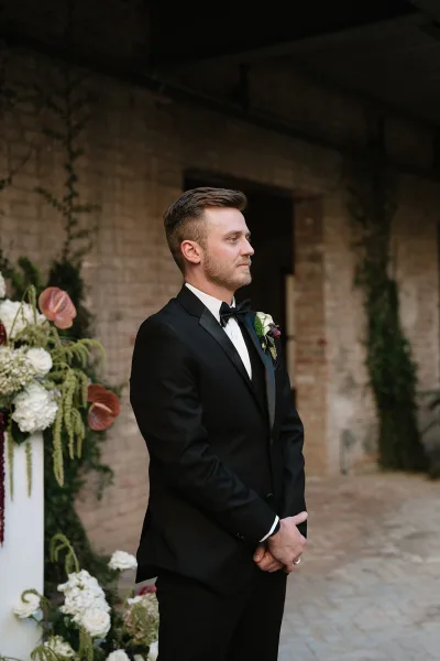 Groom portrait capturing a bow tie groom look, posed by an ivy-draped brick archway and weathered stone pavers.