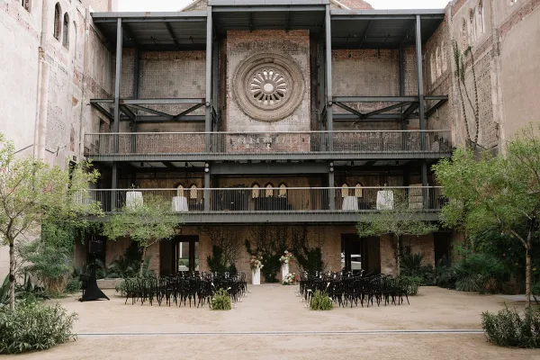 Ceremony setup with industrial ceremony decor, aisle candles in glass lining a center aisle, framed by brick walls and steel beams in a courtyard.