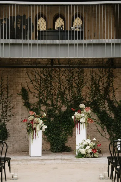Ceremony setup featuring wedding altar flowers on white pedestals against a brick wall with climbing ivy in an intimate space.
