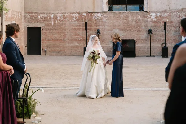 Ceremony moment with a bride holding a bouquet in an industrial courtyard against an exposed brick wall and sandy ground.