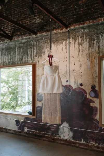 Wedding dress, a strapless mini bridal gown with a beaded overskirt, hangs against an industrial wall with a graffiti mural and a window.