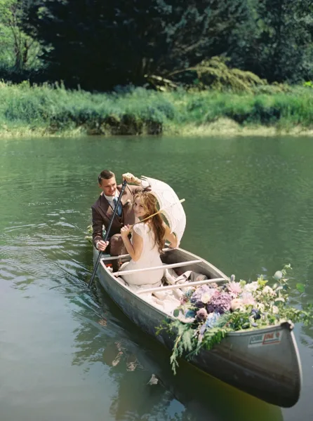 Couple portrait in a canoe wedding photo, bride with a parasol and groom paddling on a calm lake framed by trees and greenery