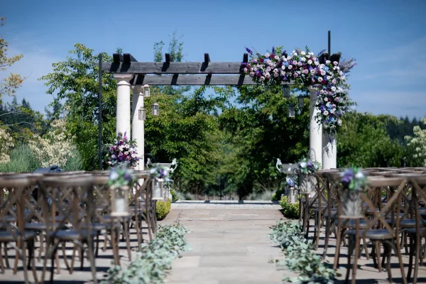 Ceremony setup with floral arch on a pergola, lanterns, and greenery-lined stone aisle with wooden chairs in a sunny garden under blue sky