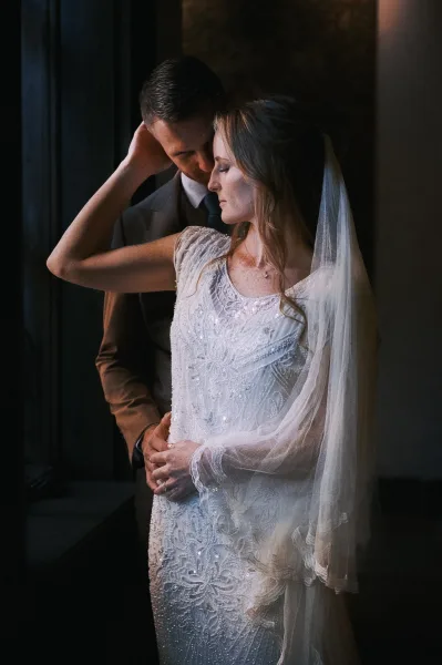 Couple portrait of bride and groom embrace, groom hugging bride as she leans in, veil draped over beaded dress in window light indoors