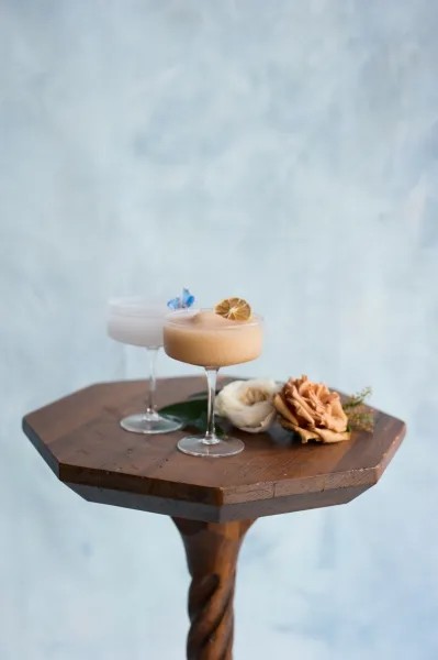 Signature cocktails in coupe glasses, frothy with citrus and edible flower accents, styled with roses on a wooden pedestal by a light blue wall