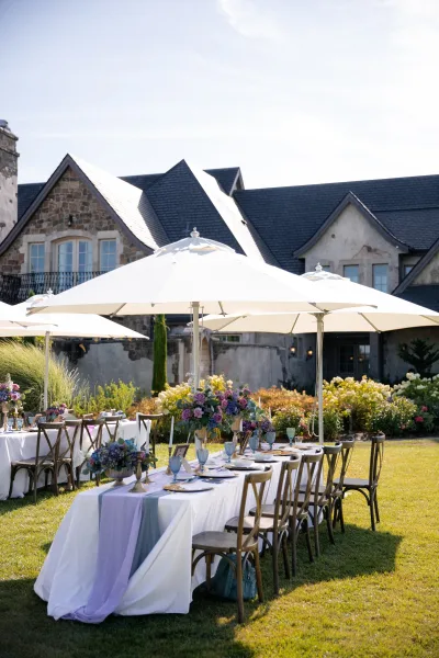 Outdoor reception tablescape with farm tables, lavender runner, floral centerpieces, taper candles and goblets beside a stone house under white umbrellas