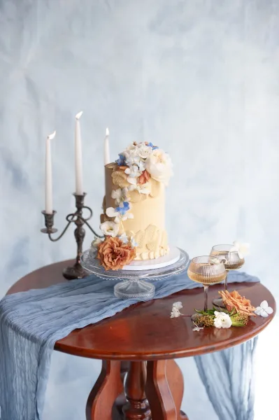 Wedding cake with buttercream wedding cake piping and a floral topper on a stand beside taper candles, coupe glasses, and a blue runner backdrop
