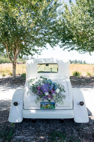 Wedding getaway car with a lush purple and blue rose-and-hydrangea arrangement on the back, parked on a gravel drive by trees and field