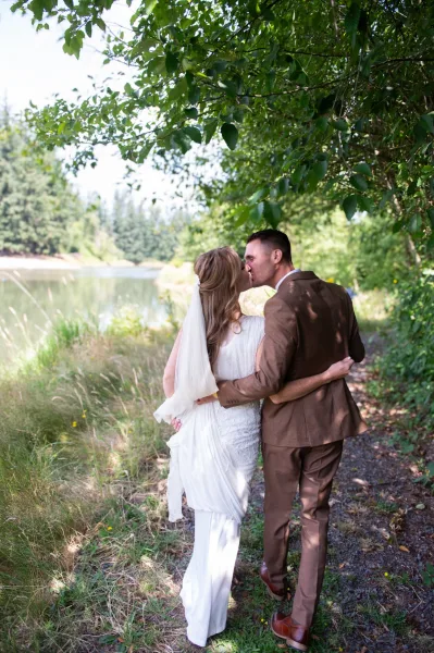 Wedding kiss portrait of bride and groom kissing from behind, her veil blowing as they walk a grassy path under trees by a lakeshore