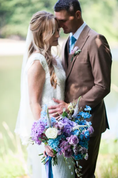 Couple portrait of bride and groom touching foreheads as she holds a blue hydrangea bouquet, veil flowing over lace dress on a green lawn