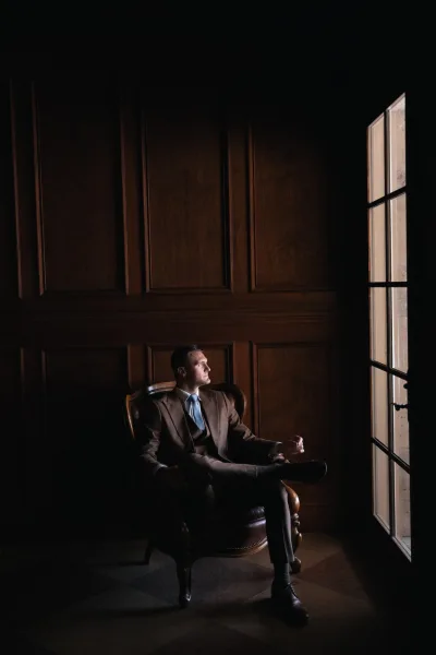 Groom portrait of a man in a three-piece suit seated in a leather armchair, lit by window light against dark wood paneling