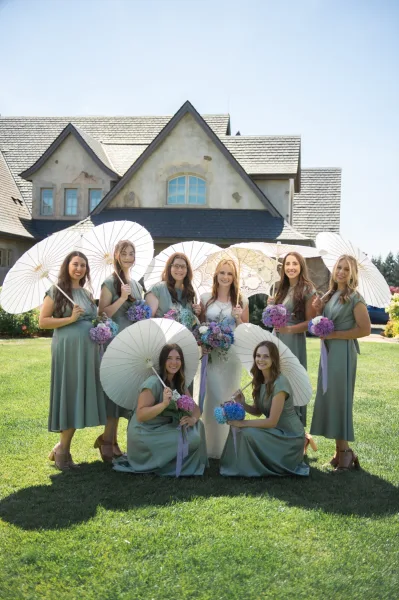 Bridesmaids group photo with the bride, holding paper parasols and pastel bouquets on an estate lawn before a large house under blue sky