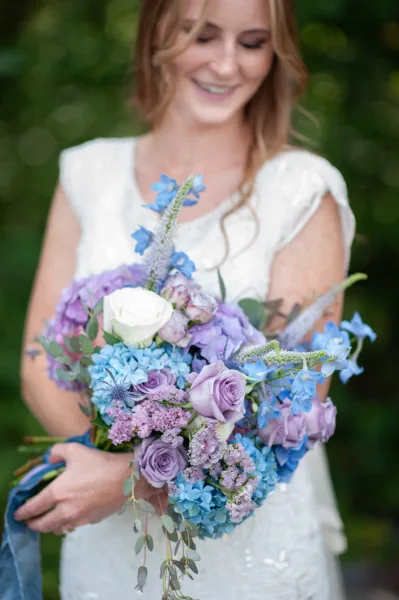 Bridal bouquet with blue and purple blooms, lavender roses, hydrangea, and eucalyptus held against a white lace wedding dress and greenery
