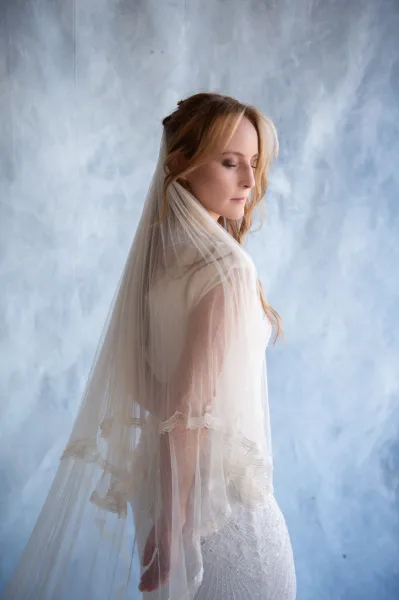 Bridal portrait of a side profile bride wearing veil and a fitted lace wedding dress against a textured blue wall, hairpiece visible