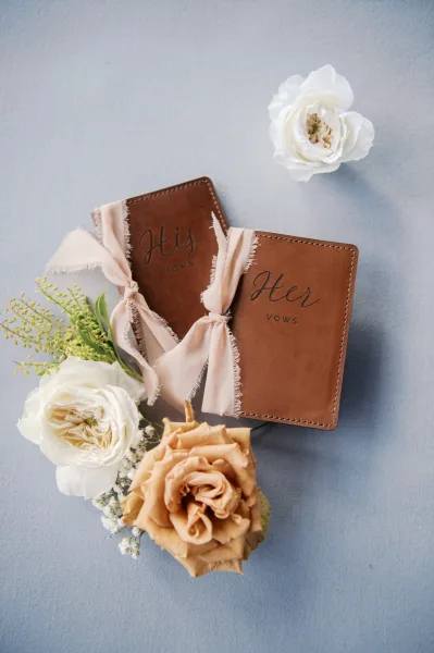 Wedding vows books with brown leather covers and blush frayed ribbon, styled with white roses and greenery on a blue fabric backdrop