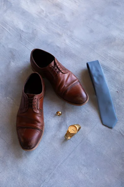 Groom details flatlay with brown leather dress shoes, light blue necktie, gold wedding band and wristwatch on a gray fabric backdrop