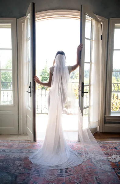 Bridal portrait of a bride from behind in a wedding dress with a long veil flowing through open French doors onto a balcony with trees beyond