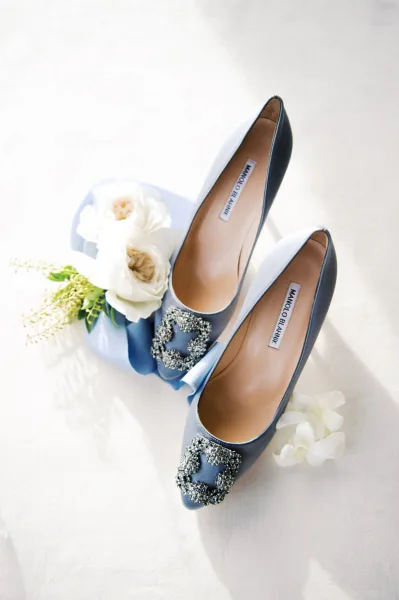 Wedding shoes in blue high heels with jeweled buckles, ribbon, and white flowers on white fabric, sunlight casting soft shadows