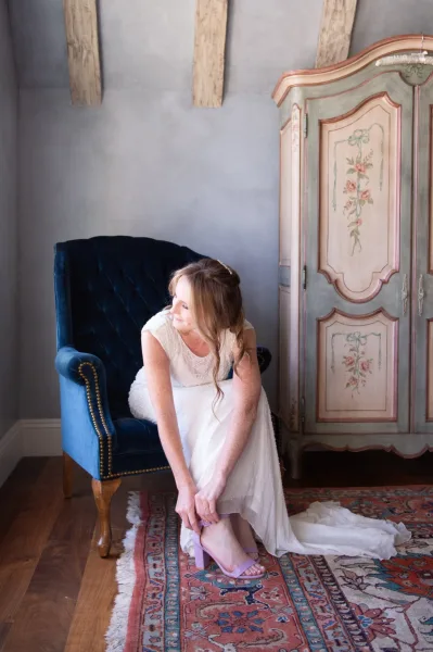 Bride getting ready putting on lavender bridal heels while seated on a tufted velvet chair, lace wedding dress train trailing on patterned rug