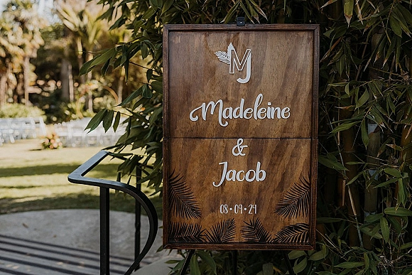 Wedding welcome sign with calligraphy lettering on a wood sign, displayed on an easel amid garden greenery and ceremony chairs outdoors