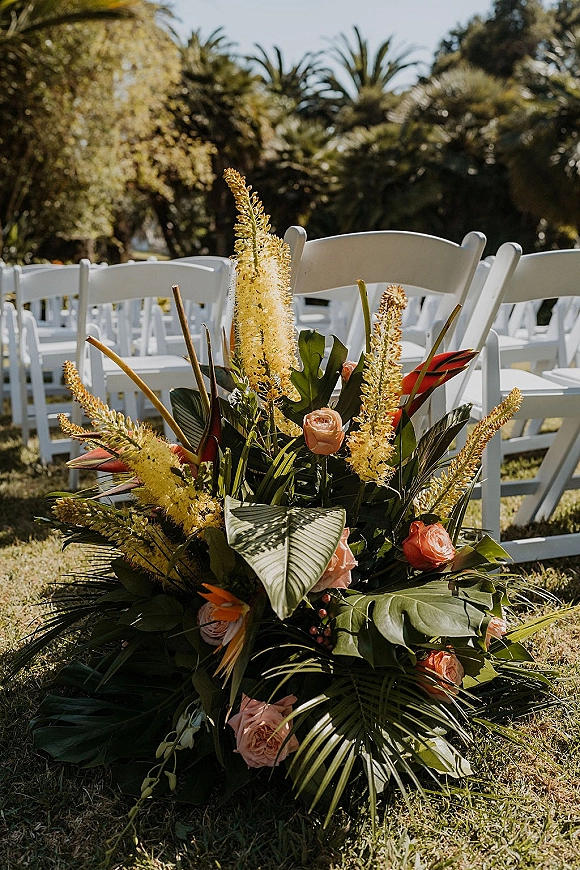 Ceremony aisle flowers with peach roses and tropical greenery lining the grass aisle beside white folding chairs in sunlit palm garden