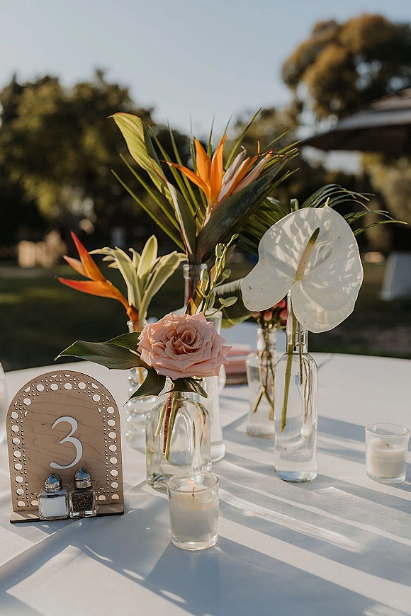 Wedding tablescape with outdoor wedding table decor, bud vases of tropical blooms and roses, votive candles, and wooden table number on a lawn
