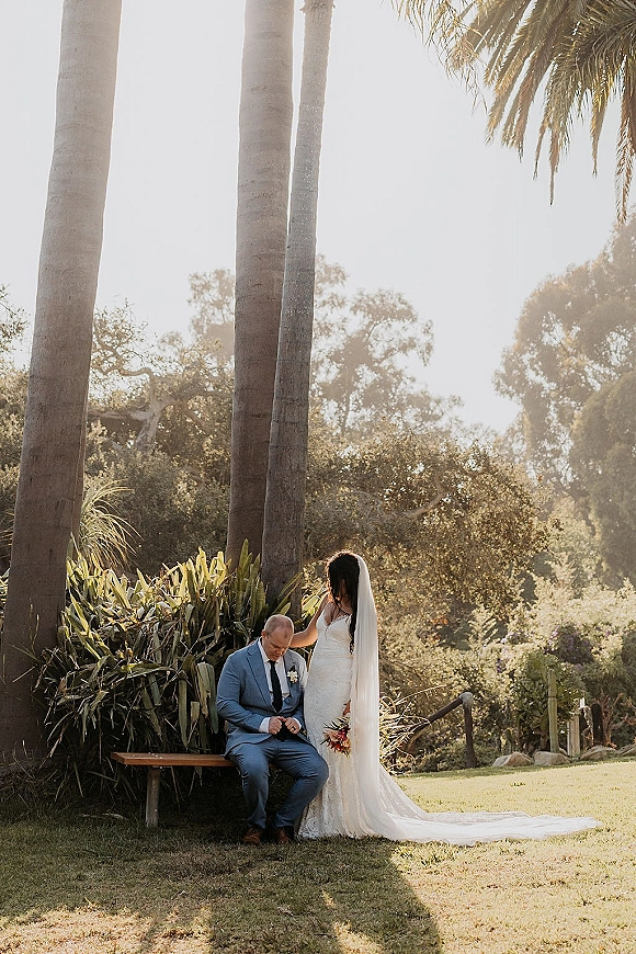 Couple portrait of bride in a lace wedding dress with cathedral veil touching groom in blue suit on a bench amid palm trees and sunlit garden greenery