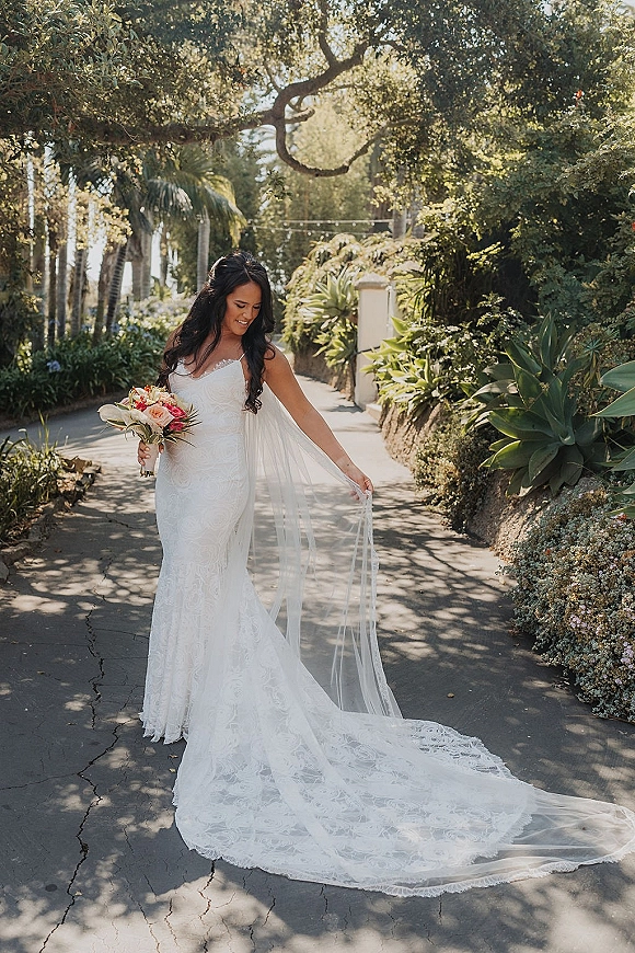 Bridal portrait of a bride holding bouquet in a lace spaghetti-strap gown with a long veil on a palm-lined garden pathway in sunlit shade