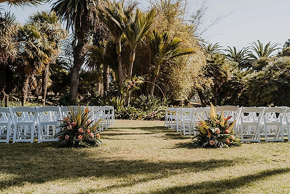Ceremony setup with white folding chairs lining an outdoor wedding ceremony aisle, accented by rose florals and tropical greenery on a lawn with palms