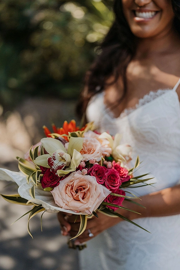 Bridal bouquet of pink and red bridal bouquet roses and orchids with greenery, held against a lace spaghetti-strap wedding dress outdoors on a tree-lined path