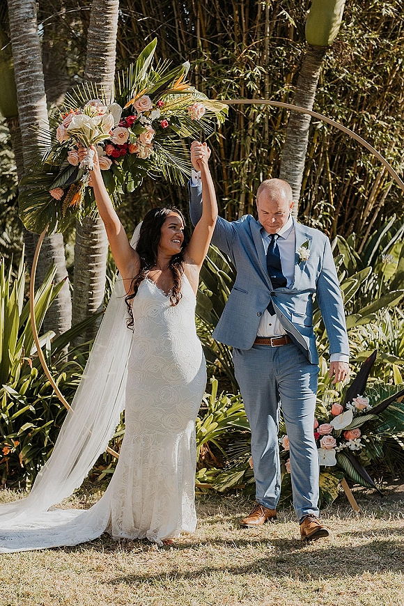 Recessional moment as bride and groom walk hand in hand, she raises a tropical wedding bouquet under a round arch in a palm garden