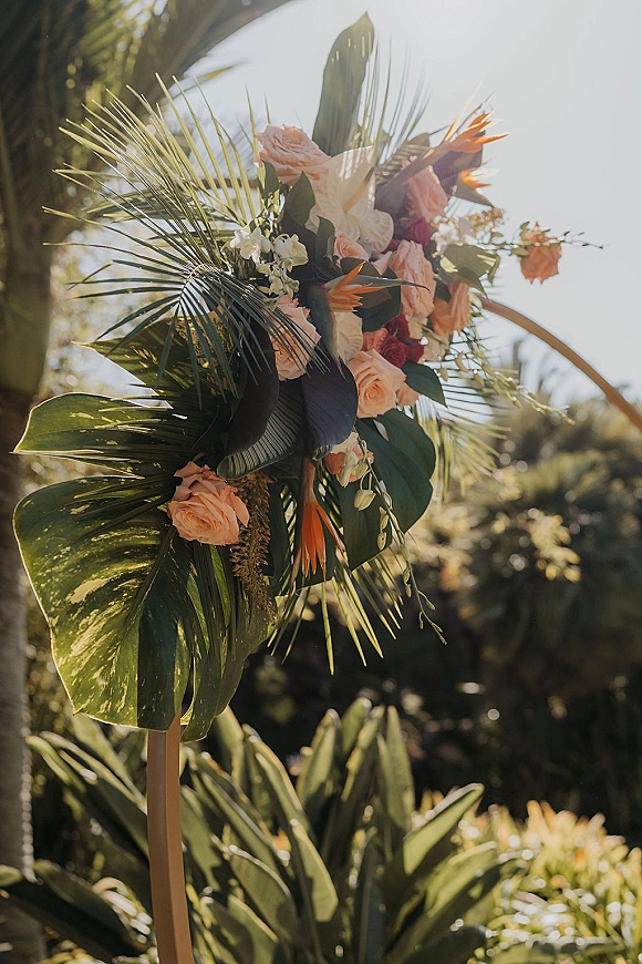 Ceremony altar flowers on a tropical wedding arch with palm fronds, monstera leaves, orchids and blush roses in a sunlit garden setting