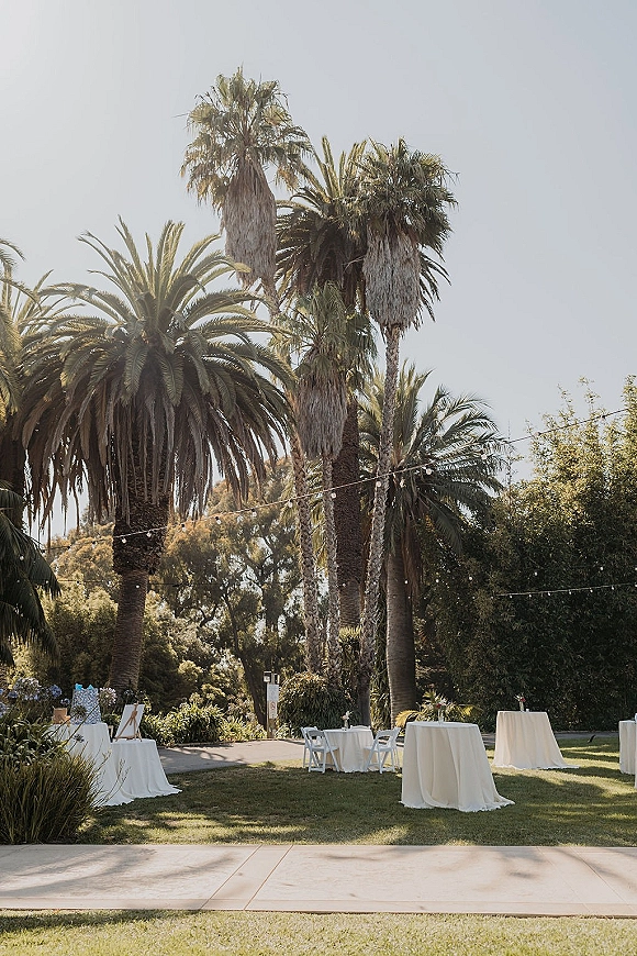 Outdoor cocktail hour setup with string lights over high top tables with white linens, bar and welcome sign on a palm-lined lawn
