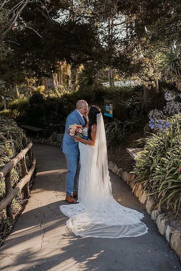 Wedding kiss portrait of bride and groom kissing on a garden path, bride in lace dress with cathedral veil holding bouquet under trees