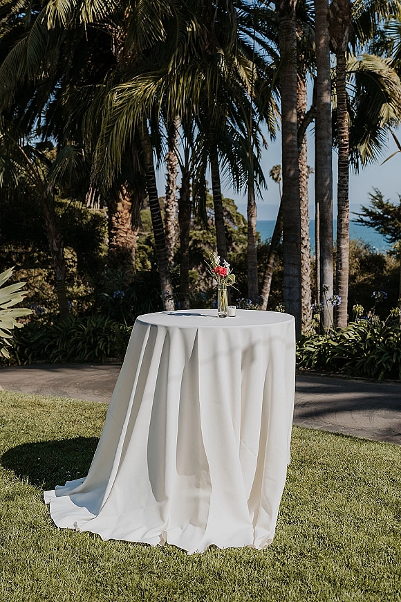 Cocktail table decor with a white linen round tablecloth, bud vase of red flowers and greenery, plus a votive candle by the ocean lawn