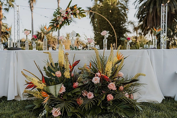 Sweetheart table decor with tropical sweetheart table florals, pink roses and bird of paradise accents beneath a floral hoop arch on a palm lawn