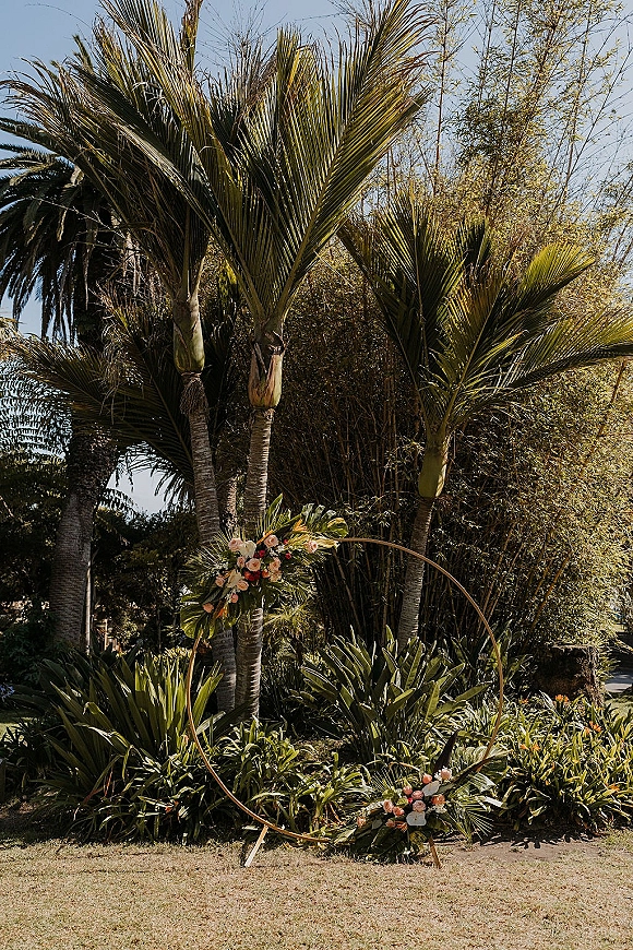 Ceremony backdrop with a circle wedding arch of greenery and roses, accented by palm fronds on a lawn with palm trees and sky