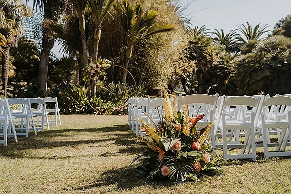 Ceremony aisle decor with a floral ground arrangement of roses and tropical greenery beside white folding chairs on a sunny lawn with palm trees