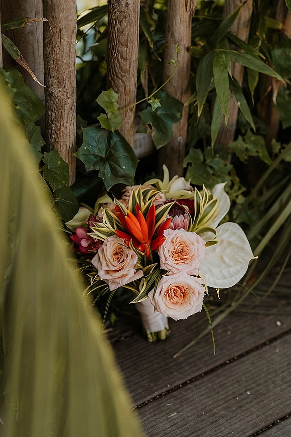 Bridal bouquet with a tropical bridal bouquet mix of blush roses, orange blooms and protea, wrapped in white ribbon on a wooden deck