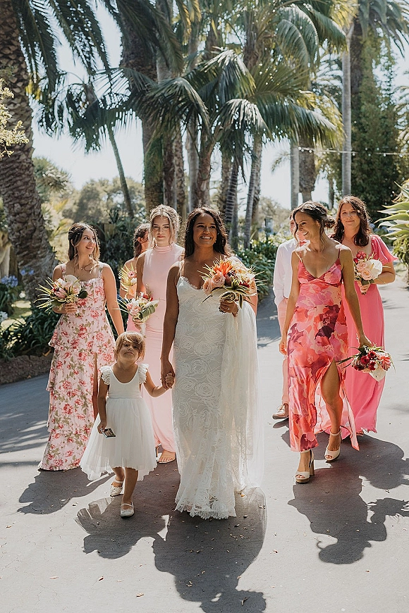 Bride with bridesmaids walking photo, bride and flower girl in lace dress with bouquets, pink dresses and palm trees along a sunny walkway