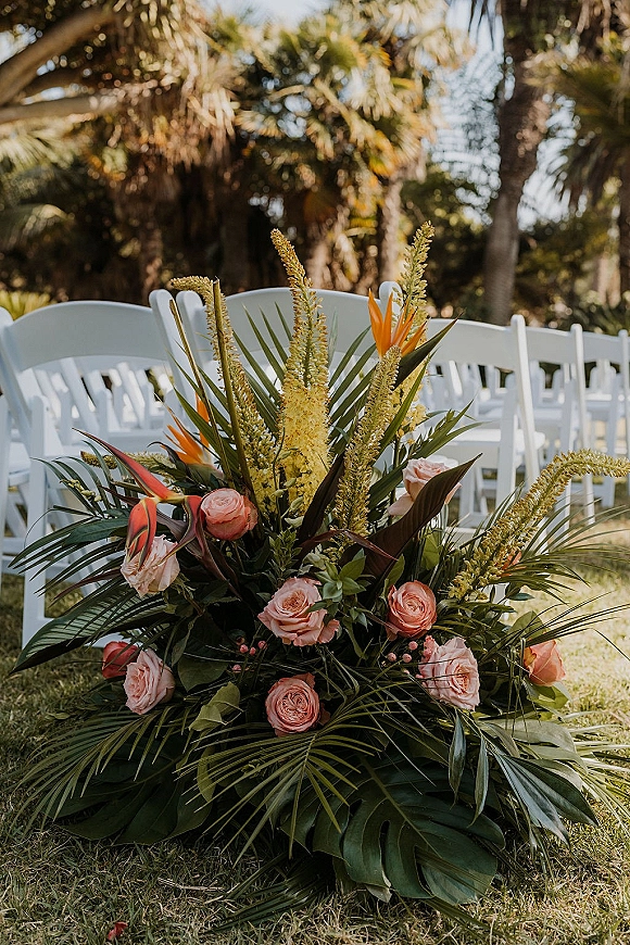Ceremony aisle flowers in a tropical aisle arrangement with pink roses, bird of paradise, and palm fronds beside white chairs on grass lawn