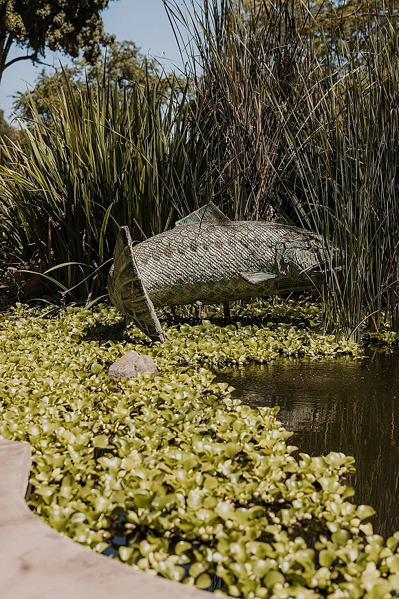 Fish sculpture outdoor fish statue rising from pond water with lily pads, framed by tall grasses, trees, and a stone ledge under sky