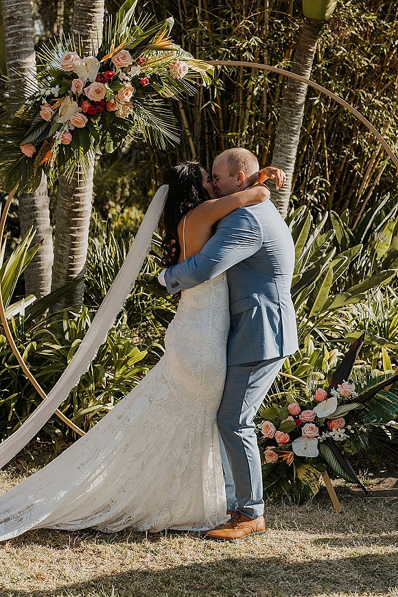 Wedding kiss under a circular wedding arch with roses, greenery, and bird of paradise, bride in lace veil and groom in suit in a tropical garden