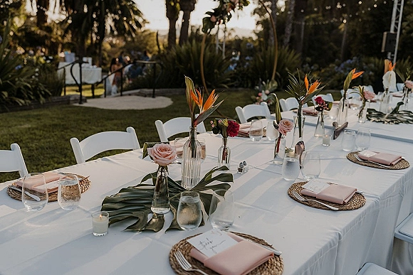 Reception tablescape with outdoor wedding table decor, blush napkins, rattan chargers, bud vases with roses and bird of paradise on a lawn