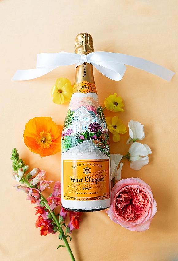 Champagne bottle with a white ribbon bow styled with pink rose and orange poppy stems on a peach backdrop flat lay