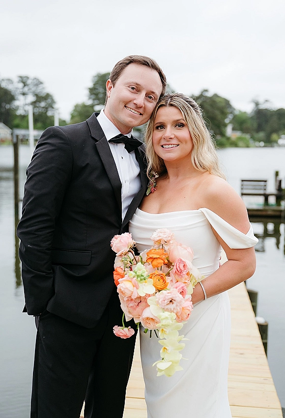Couple portrait of bride and groom portrait on a dock by a lake, bride holding bouquet in strapless dress beside groom in black tuxedo.