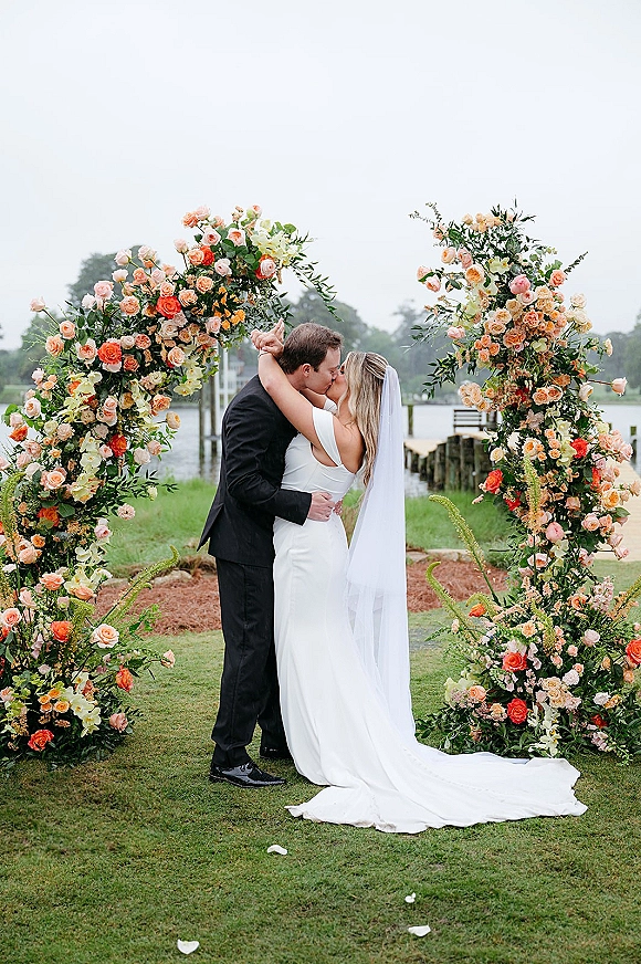 Wedding kiss portrait of bride and groom kissing under a rose and greenery floral arch, veil flowing by a waterfront dock on an overcast day