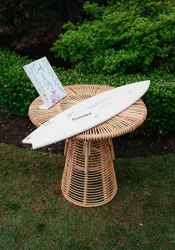 Wedding guest book surfboard guest book displayed on a wicker table with signature pen and calligraphy sign beside garden shrubs on lawn